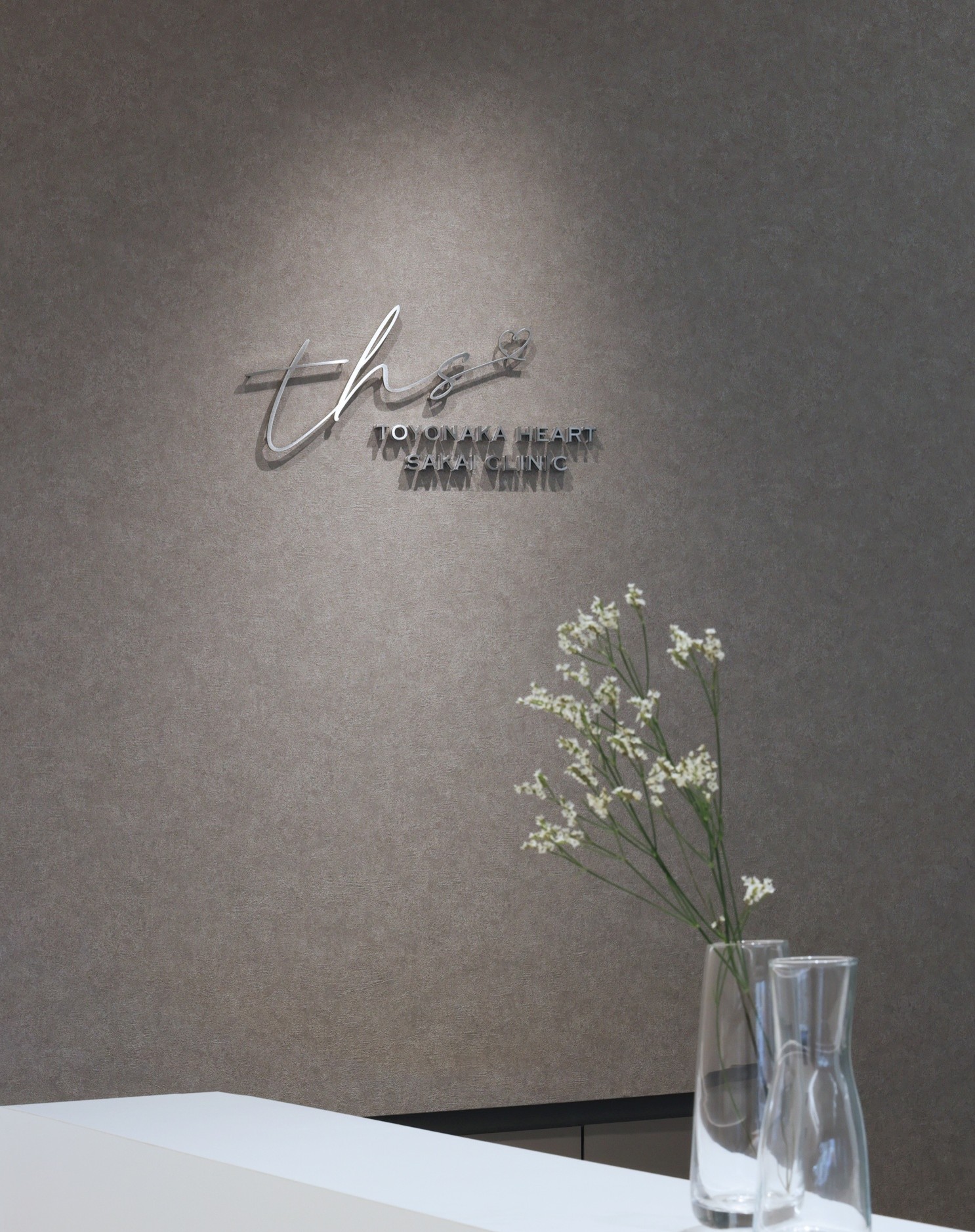 豊中駅前さかい内科・循環器クリニックの受付カウンター。上品なグレージュの壁にロゴサインが映え、ガラス花瓶に白い花が飾られた落ち着いた空間。