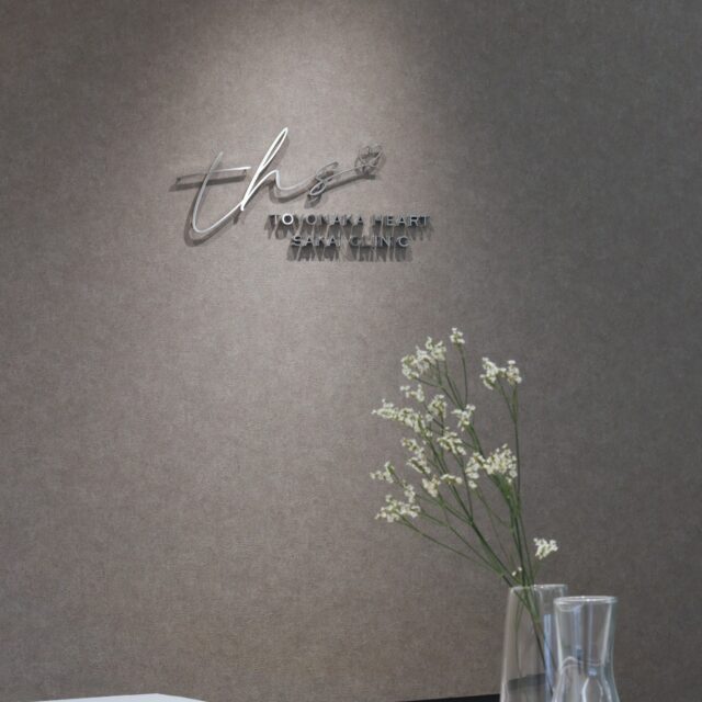 豊中駅前さかい内科・循環器クリニックの受付カウンター。上品なグレージュの壁にロゴサインが映え、ガラス花瓶に白い花が飾られた落ち着いた空間。